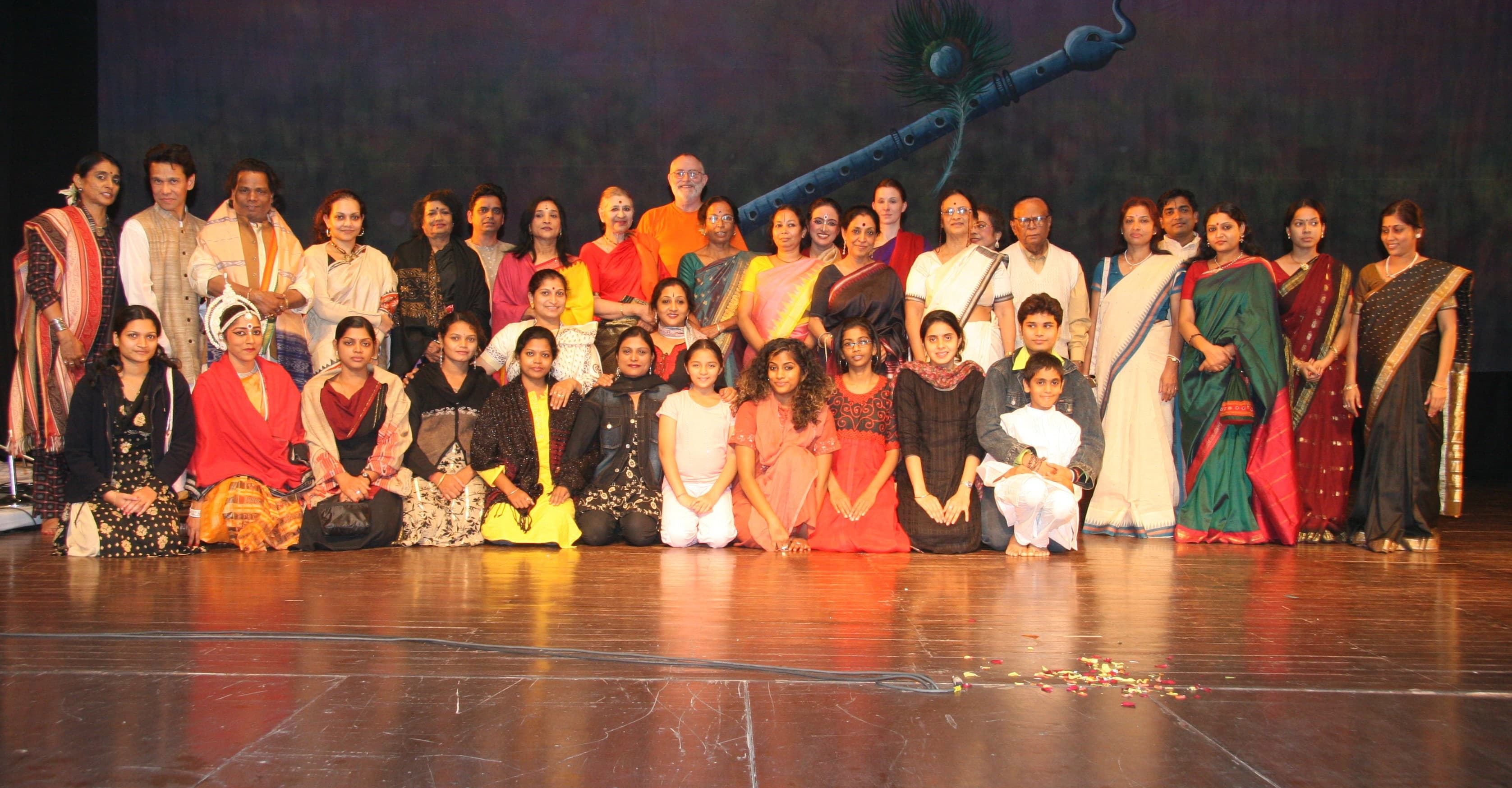 Large group of approximately 30-40 individuals, predominantly women and children, dressed in colorful traditional Indian attire, posing on stage after performance, large stylized flute or musical instrument visible against dark backdrop