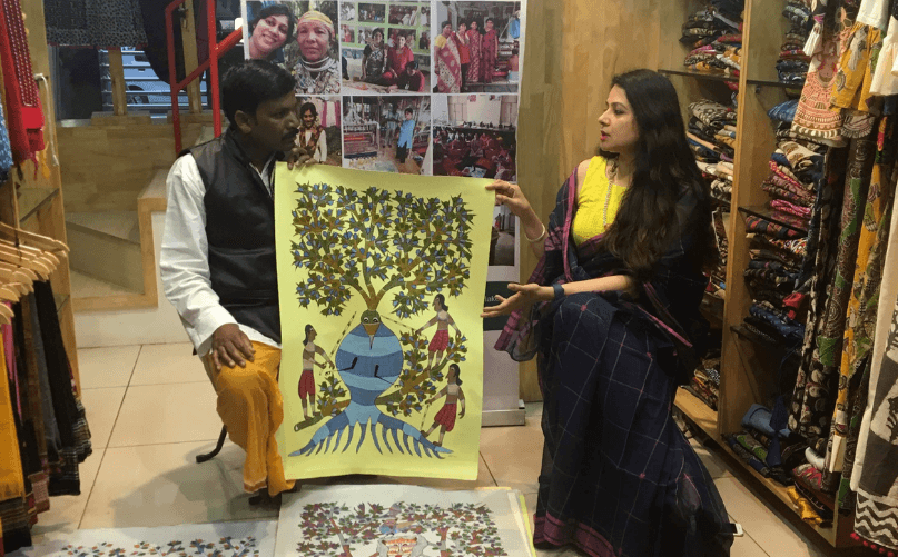 Inside retail store or exhibition space, man in white shirt and yellow dhoti seated holding up vibrant yellow painting depicting tree with human figures and blue pot at base, woman in blue and yellow saree standing next to him looking at painting, clothes racks with various fabrics visible in background