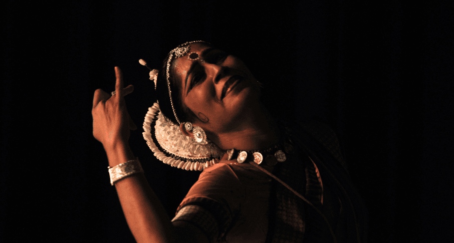 Close-up of woman performing Odissi dance, looking upwards with right arm raised in mudra, wearing elaborate traditional jewelry including large white ear ornament, necklace, and bangles, dark background with warm lighting